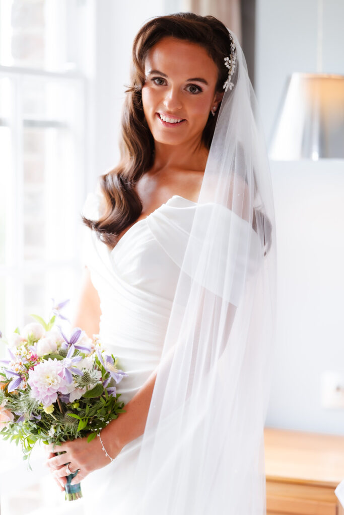 A bride in a white gown and veil stands indoors, holding a bouquet of pastel flowers. She has long brown hair and is smiling softly at the camera. Natural light comes through a nearby window.