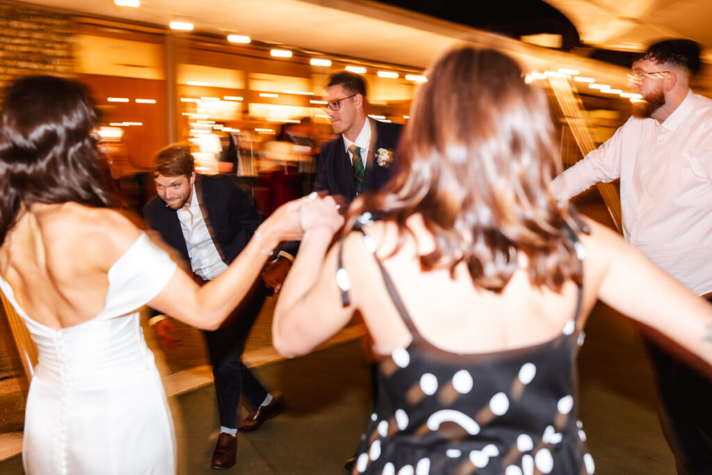 A group of people, dressed in formal attire, hold hands and dance in a circle at an outdoor nighttime event with warm, blurred lights in the background. The image captures a lively, joyful moment in motion.