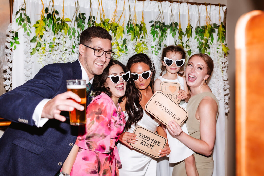 Five people pose for a festive photo booth picture at a wedding, wearing heart-shaped sunglasses and holding signs that say I ❤️ YOU, #TEAMBRIDE, and TIED THE KNOT. One person holds a drink. Greenery decorates the background.