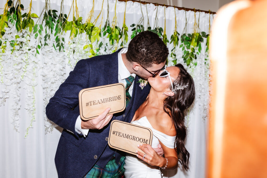 A bride and groom kiss while holding wooden signs reading “#TEAMBRIDE” and “#TEAMGROOM” in front of a backdrop with greenery and white flowers.