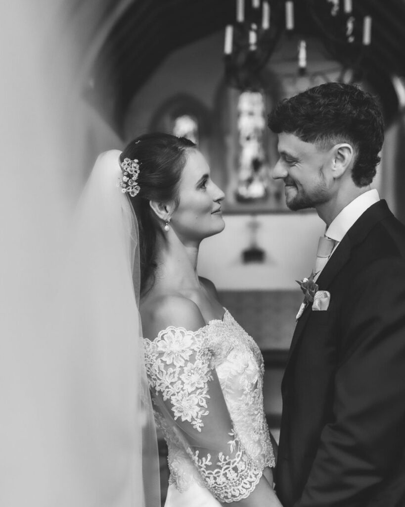 A bride and groom stand facing each other, smiling, inside a church. The bride wears an off-shoulder lace gown and veil; the groom is in a suit with a boutonniere. Stained glass windows appear in the background.