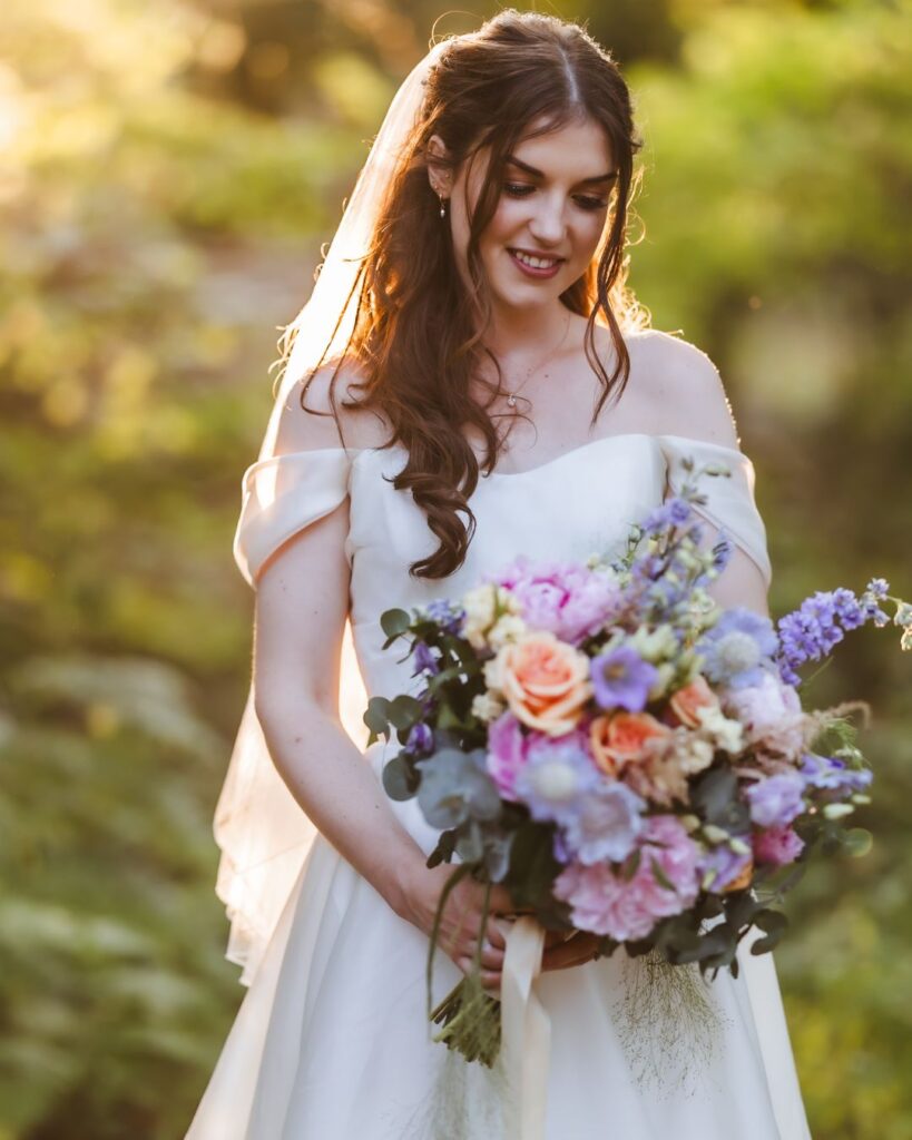 A bride with long brown hair in a white off-shoulder wedding dress smiles while holding a colorful bouquet, captured by a Hampshire wedding photographer outdoors in soft, golden sunlight.