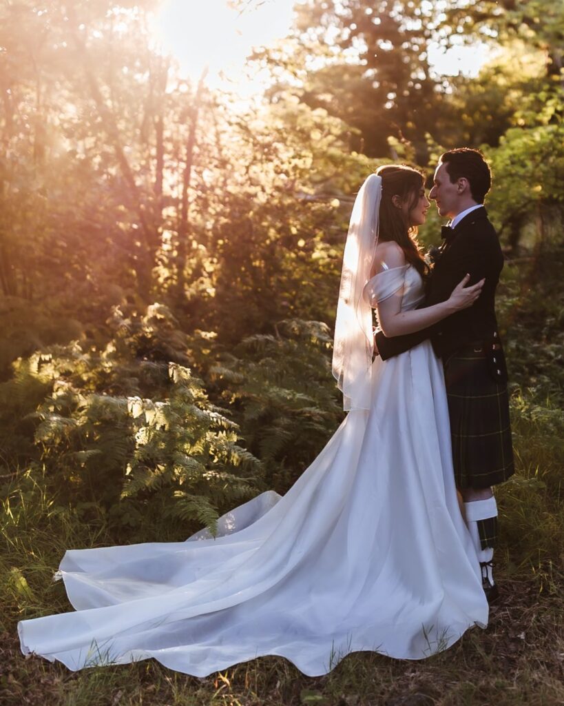 A bride in a white gown and veil embraces her groom in a dark jacket and kilt in a sunlit forest, surrounded by greenery and soft, golden light—captured beautifully by a Hampshire wedding photographer.