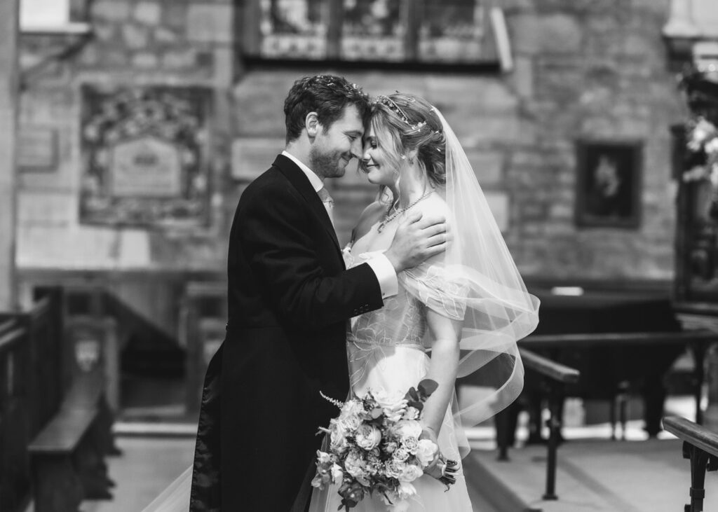 A bride and groom stand in an embrace inside a church, gazing into each other’s eyes and smiling. Captured in black and white by a Hampshire wedding photographer, the bride holds a bouquet and wears a veil, while the groom is in a suit.
