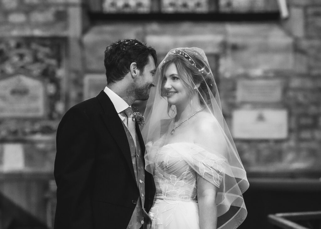 A bride and groom stand close indoors, the bride smiling in a veil and strapless gown as the groom in a suit gazes at her; captured by a Hampshire wedding photographer, the background features blurred stone walls and plaques.