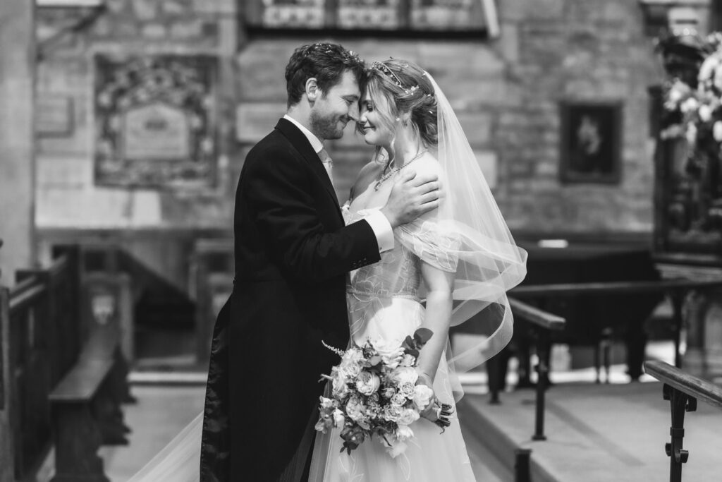 A bride and groom stand close together in a church, smiling lovingly at each other. Captured by a Hampshire wedding photographer, the intimate black-and-white scene highlights their romantic connection, with the bride in her veil and gown holding a bouquet.