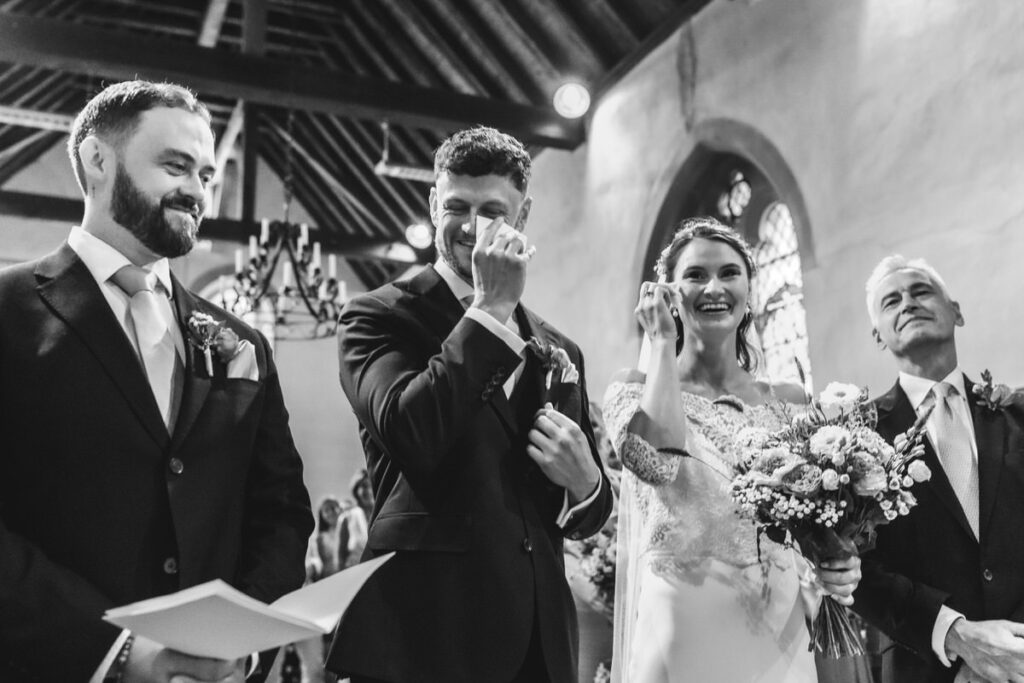 A black and white photo of a joyful wedding ceremony shows the groom wiping away tears, the bride smiling and holding a bouquet, and two men in suits standing beside them, all inside a church with high wooden ceilings.