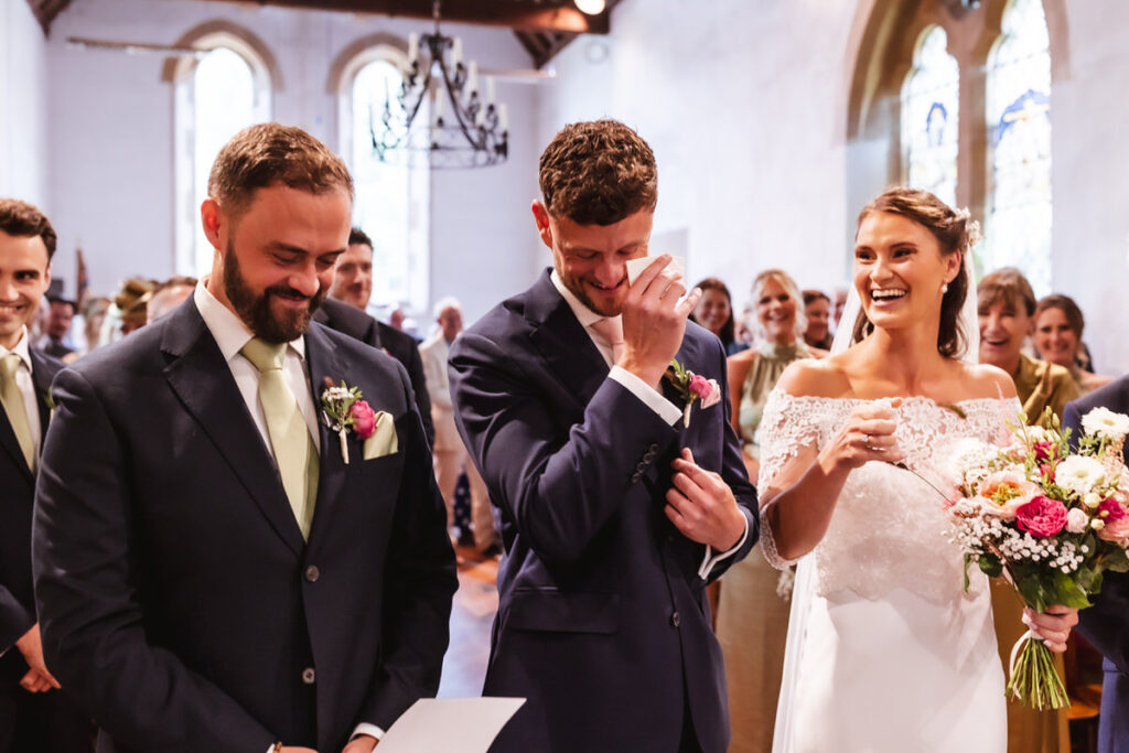 A groom wipes a tear while standing with a smiling bride and best man at a wedding ceremony inside a church, surrounded by happy guests.