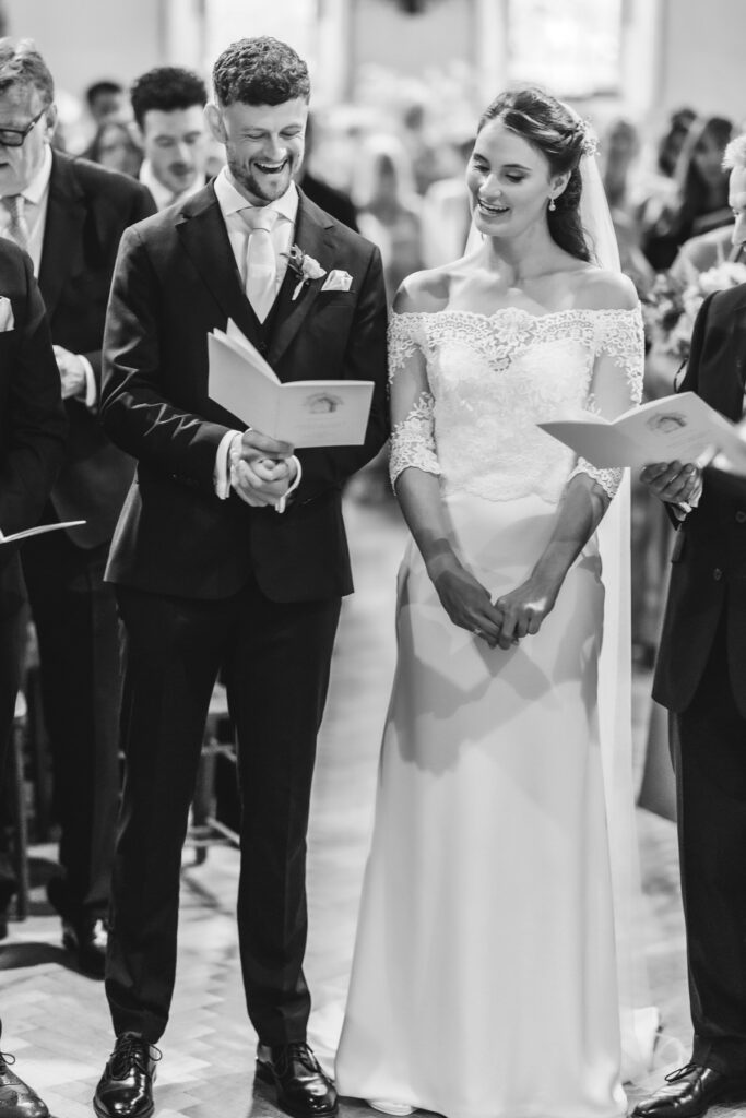 A bride and groom stand side by side during a wedding ceremony, smiling and holding programs. The groom wears a dark suit and the bride wears an off-the-shoulder lace wedding dress. Guests stand around them in a church.