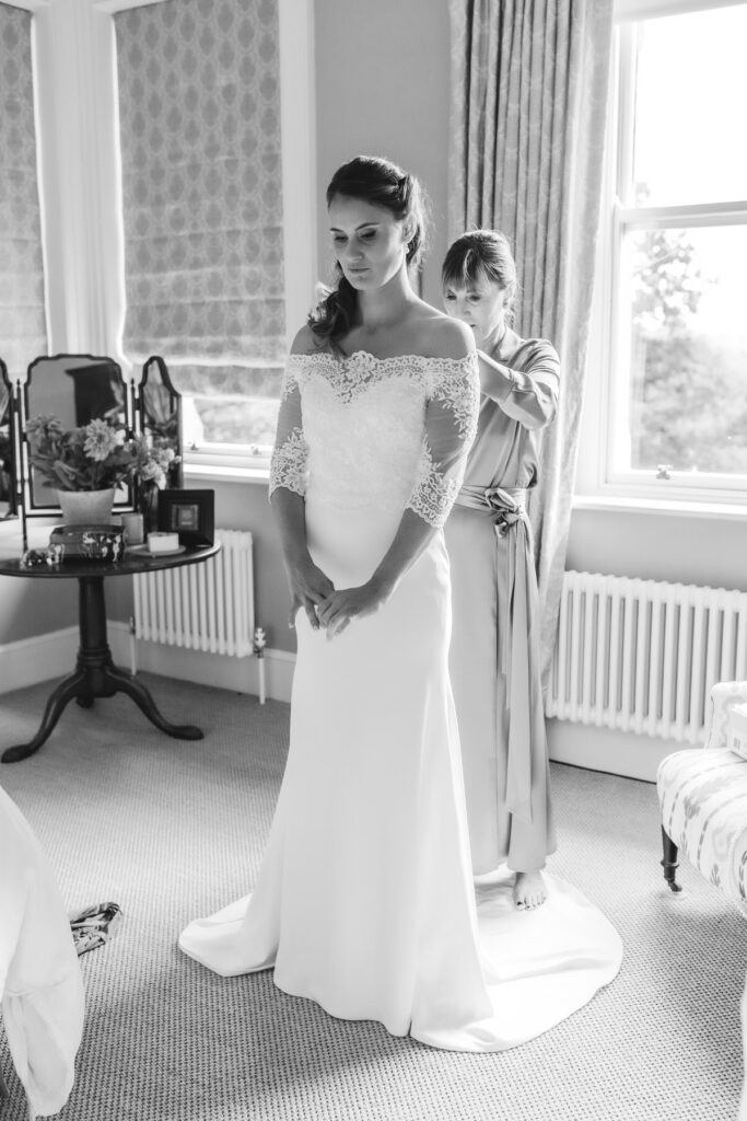 A bride in an off-the-shoulder lace wedding dress stands still while another woman helps adjust her dress in a bright, elegant room with large windows and a dressing table.