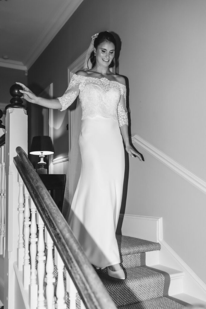 A woman in a white lace wedding dress and veil smiles as she walks down carpeted stairs in a well-lit home interior.