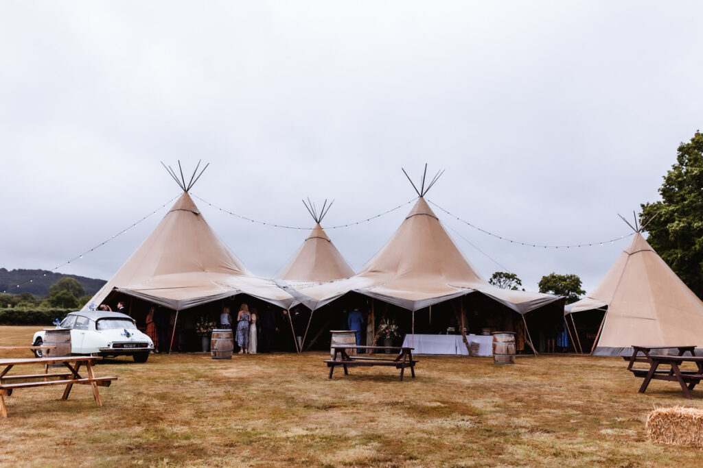 Large cream tipi tents are set up outdoors on a grassy field, with picnic tables and hay bales nearby. A classic white car is parked on the left. String lights are draped between the tents under a cloudy sky.