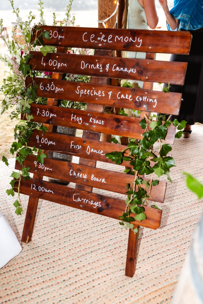 A wooden sign decorated with green ivy lists a wedding schedule in white writing, including ceremony, drinks, speeches, hog roast, first dance, cheese board, and carriages with corresponding times.