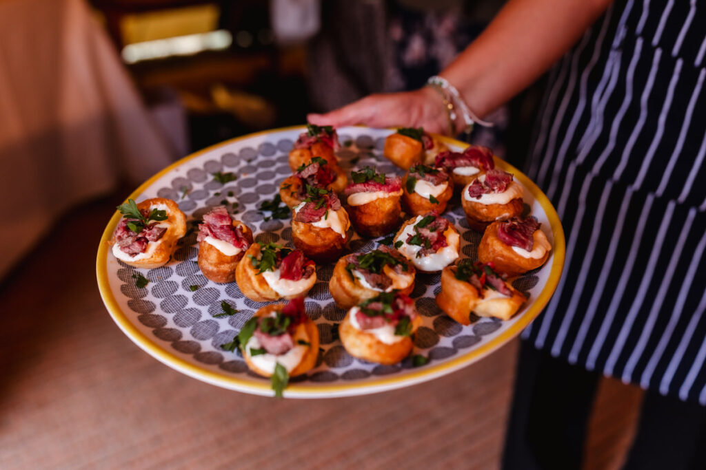 A person wearing a striped apron holds a large plate of small, round appetizers topped with meat, sauce, and fresh herbs. The platter is patterned with dots and the background is softly blurred.