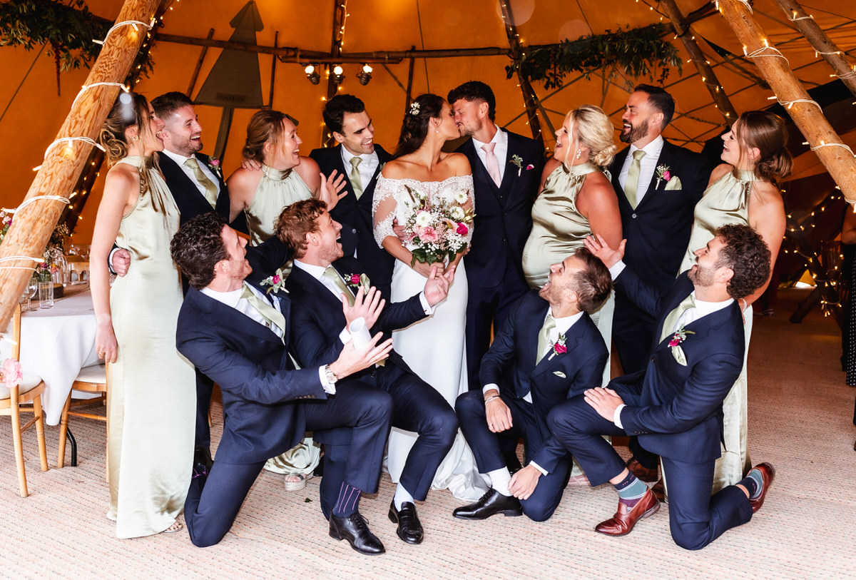 A bride and groom kiss in the center of their wedding party, surrounded by bridesmaids in sage green dresses and groomsmen in navy suits, all smiling and celebrating inside a decorated tent.