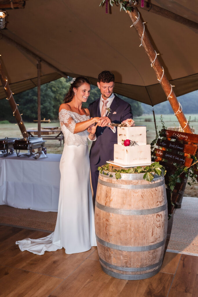 A bride and groom smile as they cut a white wedding cake together on a rustic wooden barrel inside a tent, with fairy lights and a scenic outdoor background visible.