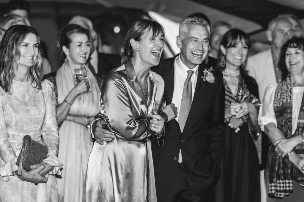 A group of well-dressed people, including a smiling couple in the center, stand closely together laughing and holding drinks at what appears to be a celebratory event. The photo is in black and white.