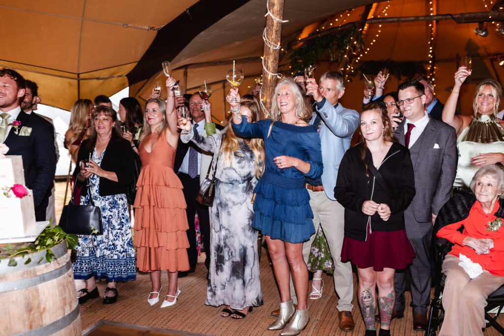 A group of people at a wedding celebration raise their glasses in a toast, smiling and dressed in colorful semi-formal attire, standing inside a tent decorated with string lights.