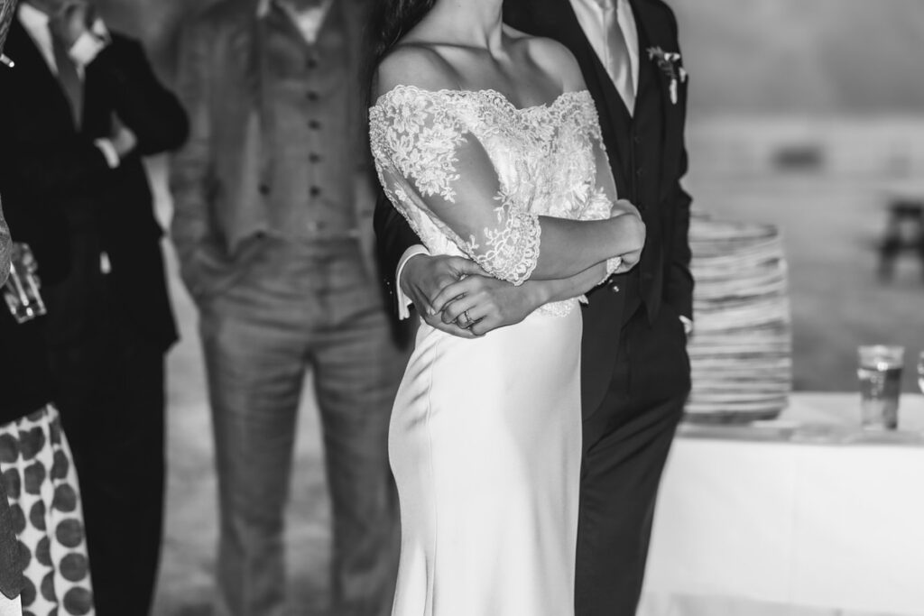 A black and white photo of a bride in an off-the-shoulder lace wedding dress with a groom standing behind her, his arm around her waist, surrounded by other people dressed formally.