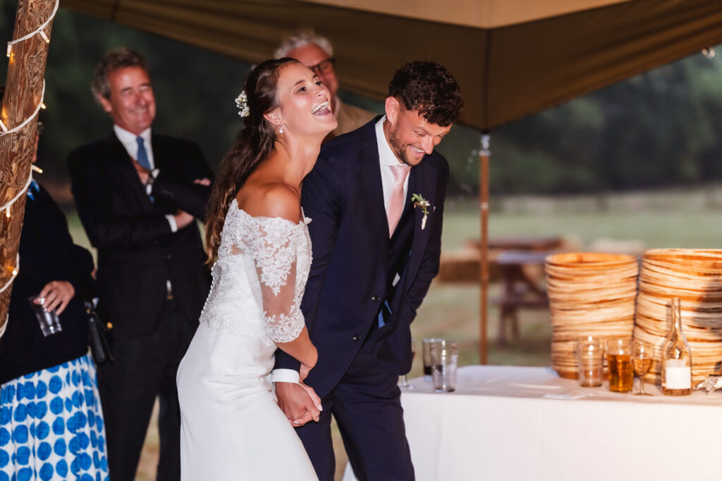 A bride and groom, both smiling and laughing, hold hands at their wedding reception under a tent. Guests in formal attire stand behind them, and a table with drinks and stacked baskets is nearby.