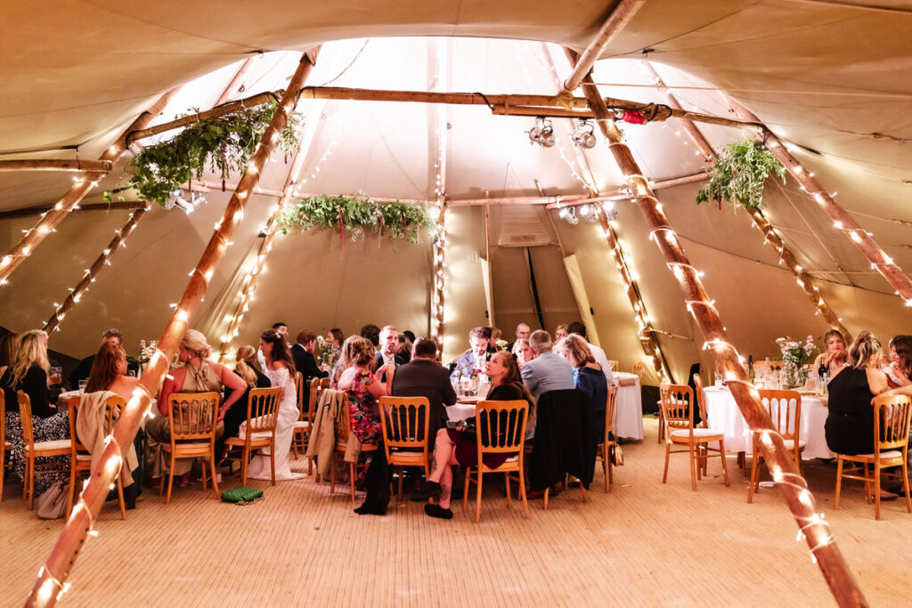 Guests sit around decorated tables inside a large, warmly lit tent with wooden beams wrapped in string lights and greenery hanging from the ceiling, creating a festive atmosphere.