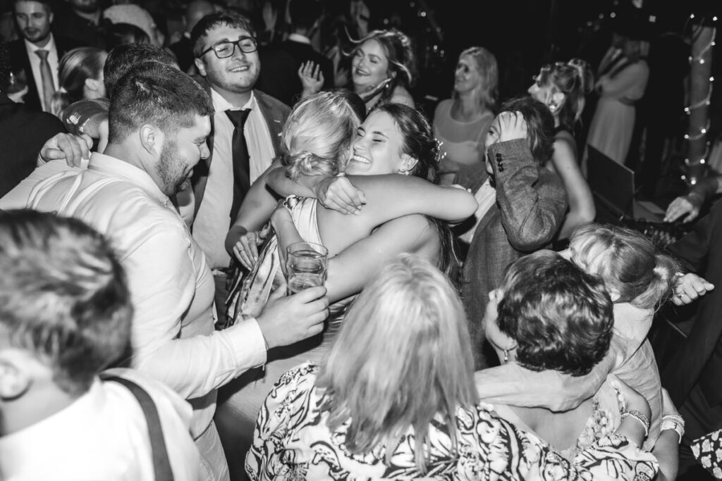 A group of people at a lively party or celebration. Two women in the center share a joyful hug, surrounded by smiling and laughing guests. The image is black and white.