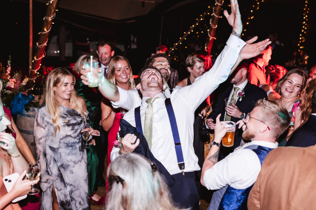 A group of people dressed in formal wear dance and celebrate at a lively party, with one man in the center raising his arms and smiling joyfully while others laugh and hold drinks around him.