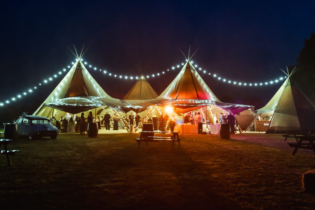 A large outdoor tent setup with three connected tipis, glowing with warm lights at night. String lights hang above, and people gather inside. A vintage car and picnic tables are visible on the grassy area in front.