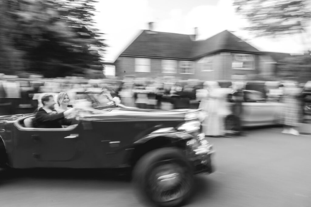A blurred black-and-white photo by a Hampshire wedding photographer captures a couple driving a vintage convertible past onlookers outside residential houses, creating a sense of motion and excitement.