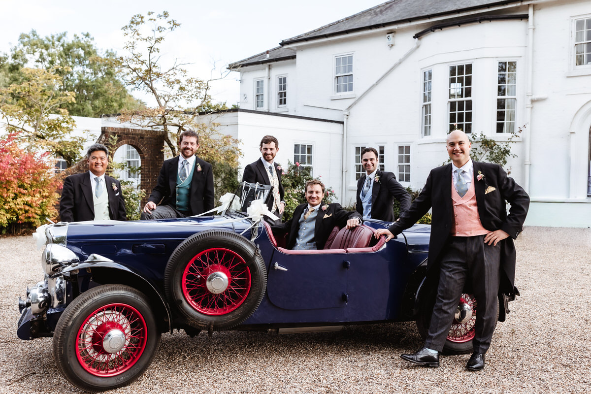 Six men dressed in suits pose around a vintage navy blue car with red wheels, parked before a grand white building—an unforgettable moment captured by a Hampshire wedding photographer amid lush greenery and arched windows.