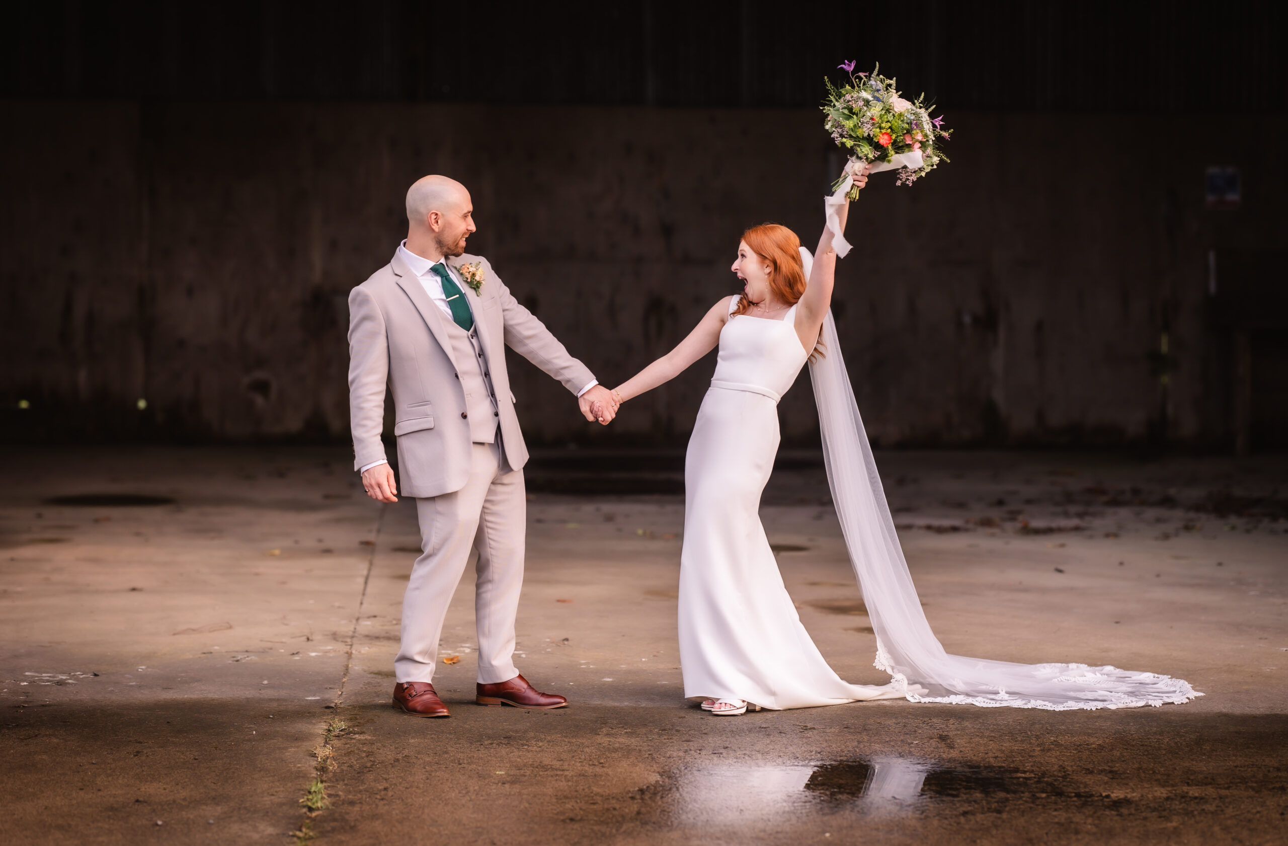 A bride and groom stand on a concrete floor, holding hands and smiling at each other. The bride raises her bouquet victoriously, and her veil trails behind her. The groom wears a light suit and the background is dark and industrial.