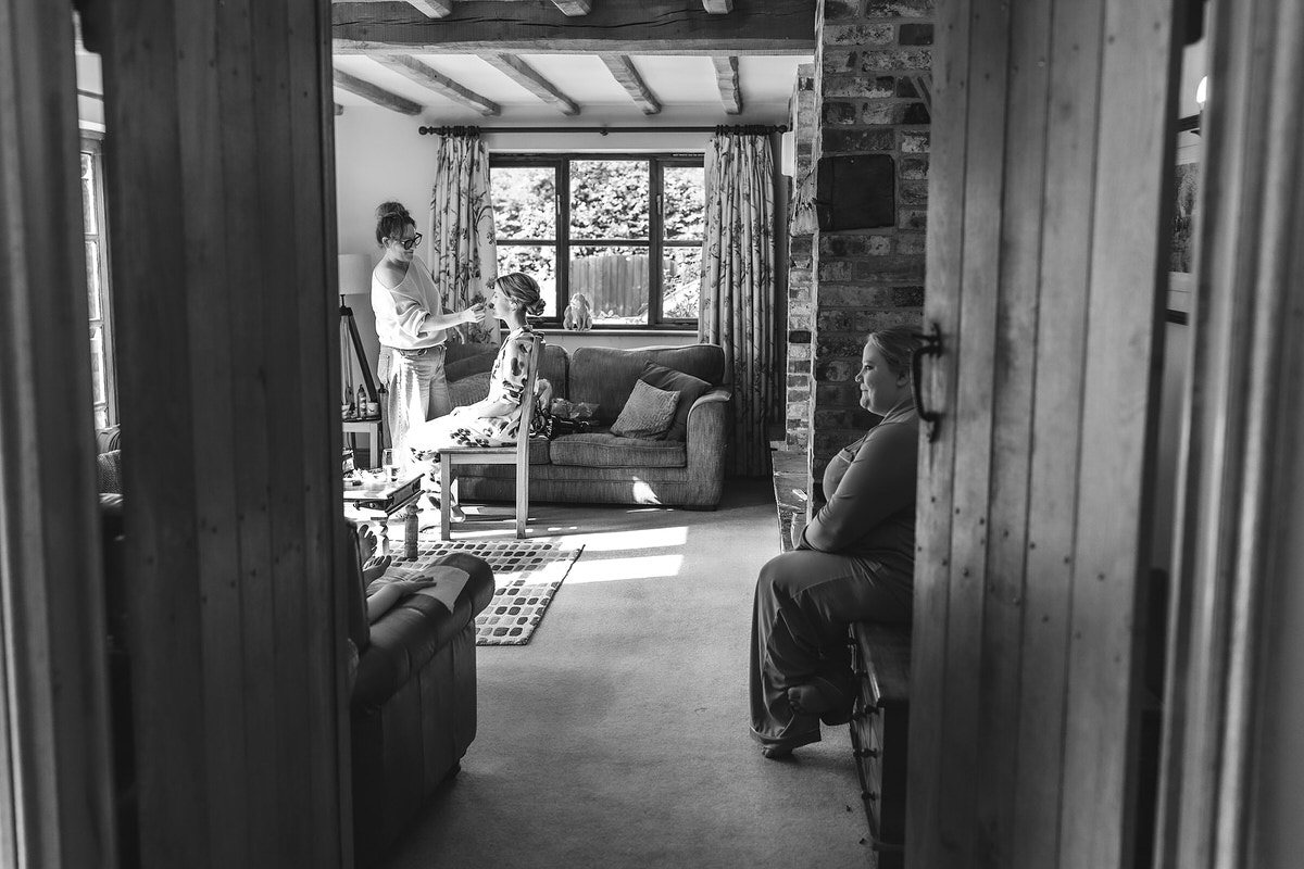 Black and white photo of a cozy living room. A woman styles a childs hair on a sofa near a window, while another woman sits on a bench in an adjoining hallway, watching the scene.