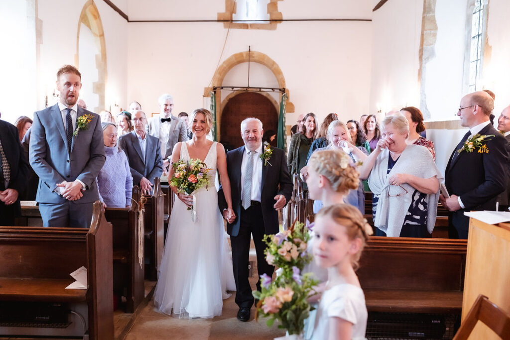 A bride holding a bouquet walks down the aisle with an older man in a church, smiling guests watching and flower girls in front. The groom stands on the left, looking surprised.
