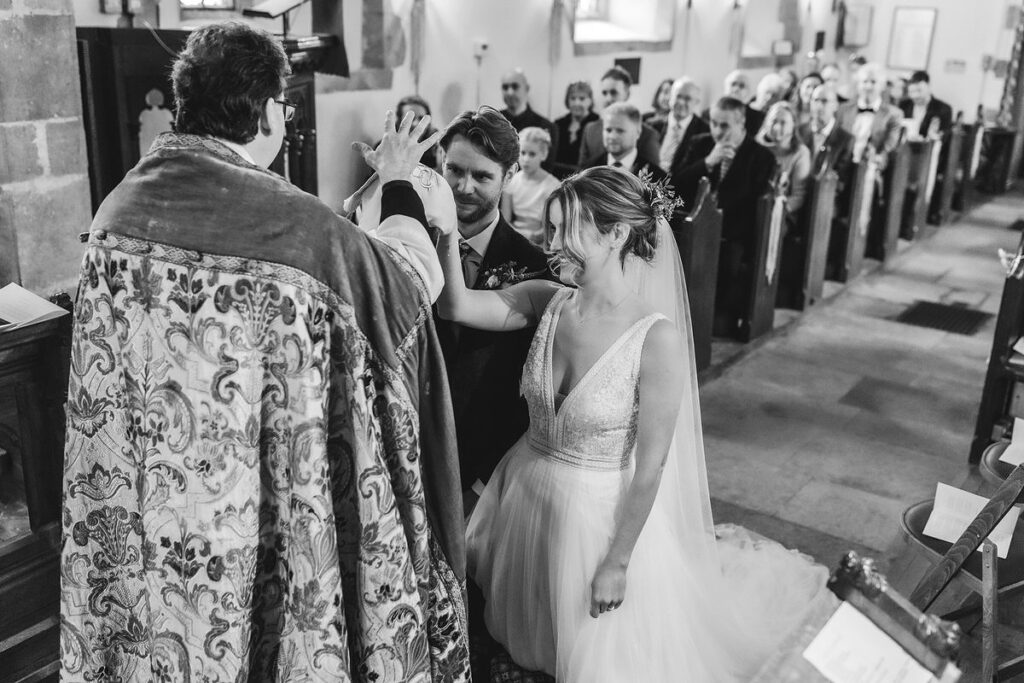 A bride and groom stand at the altar in a church, smiling as the officiant gestures in blessing. Guests watch from the pews. The scene is captured in black and white.