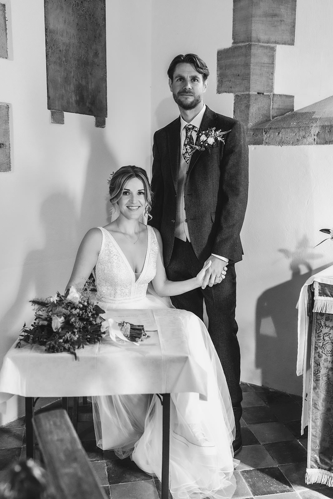 A bride and groom pose indoors; the bride sits at a small table holding a bouquet, while the groom stands beside her, holding her hand. Both are smiling gently, dressed in wedding attire.