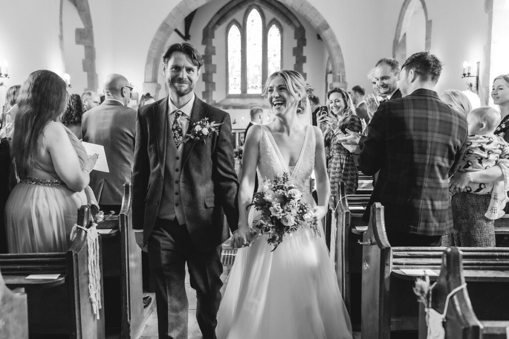 A smiling bride and groom walk down the aisle together in a church, surrounded by cheering guests. The bride holds a bouquet and wears a wedding dress; the groom wears a suit. The image is in black and white.