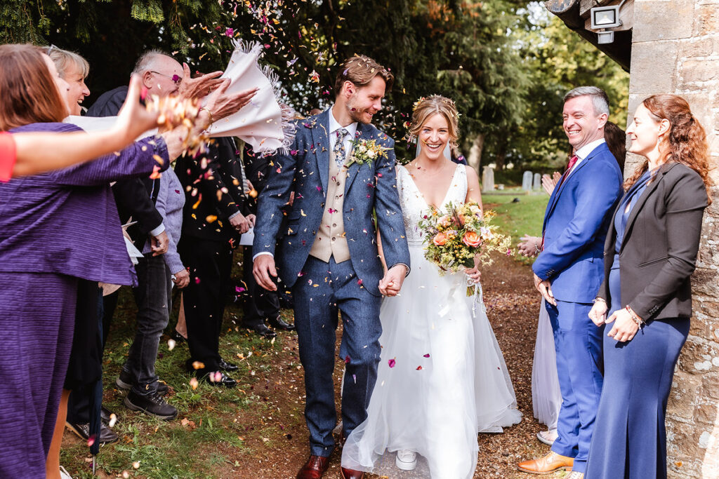 A bride and groom smile as they walk hand in hand outdoors, surrounded by cheering guests who throw confetti over them. The groom wears a blue suit and the bride wears a white dress, holding a bouquet.