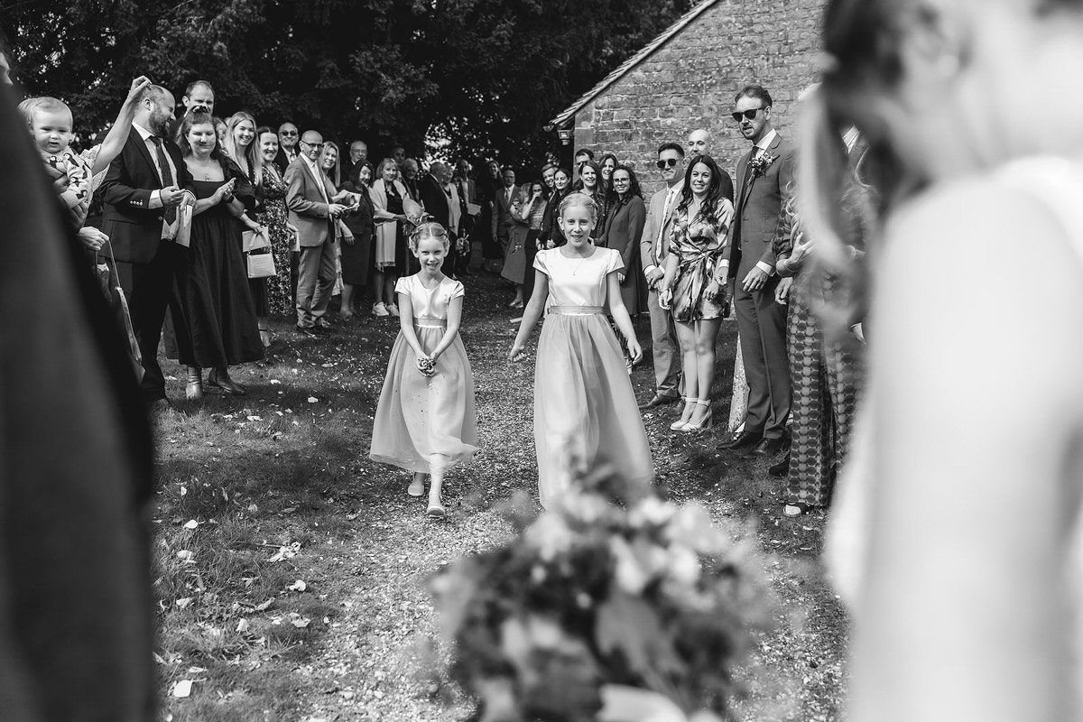 Two young flower girls in matching dresses walk down an outdoor aisle, smiling, as guests stand on either side watching. The scene is in black and white, with trees and a brick building in the background.
