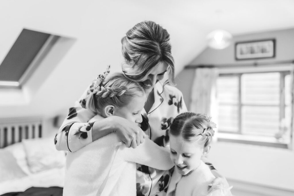 A woman in a floral robe lovingly hugs two young girls with braided hair in a bright, cozy bedroom. The tender moment is filled with warmth and joy.