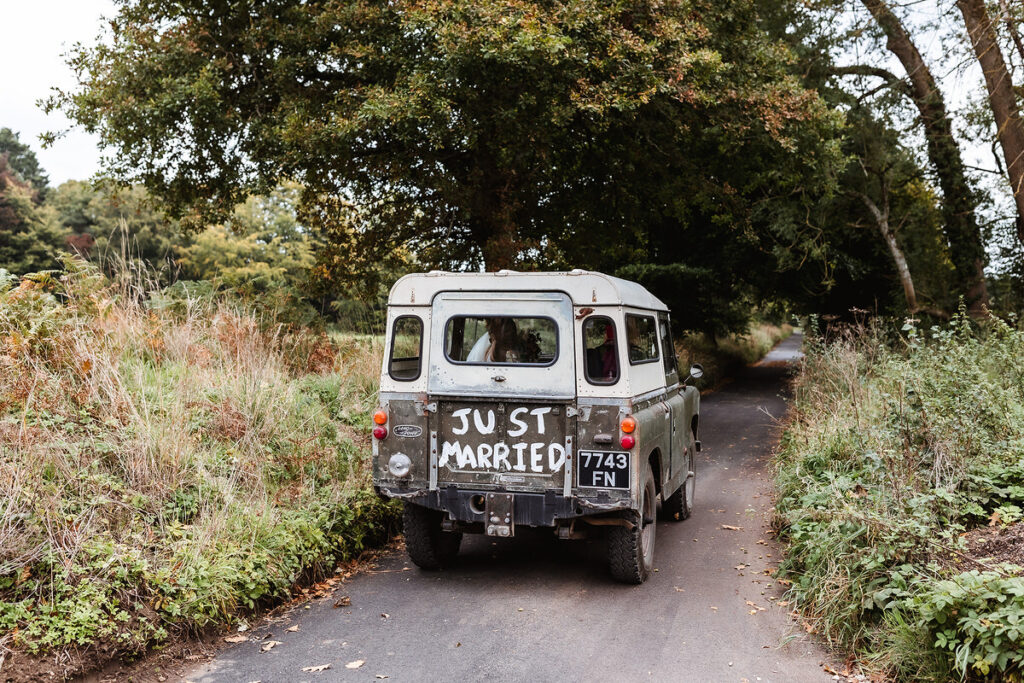 A vintage SUV with JUST MARRIED painted on the back window drives down a narrow, tree-lined country road surrounded by greenery.