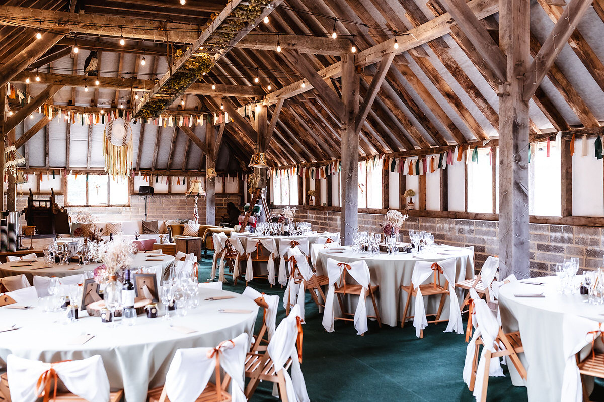A rustic barn decorated for a wedding reception, with round tables covered in white cloths, wooden chairs with white sashes, string lights, and dried floral centerpieces under a high wooden-beam ceiling.
