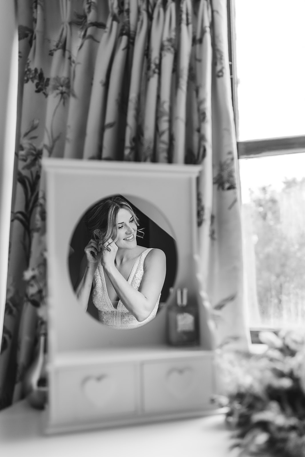 A woman in a white dress is reflected in a mirror as she smiles and puts on an earring. She is in front of patterned curtains and a window, with a vanity table visible in the foreground. The image is in black and white.