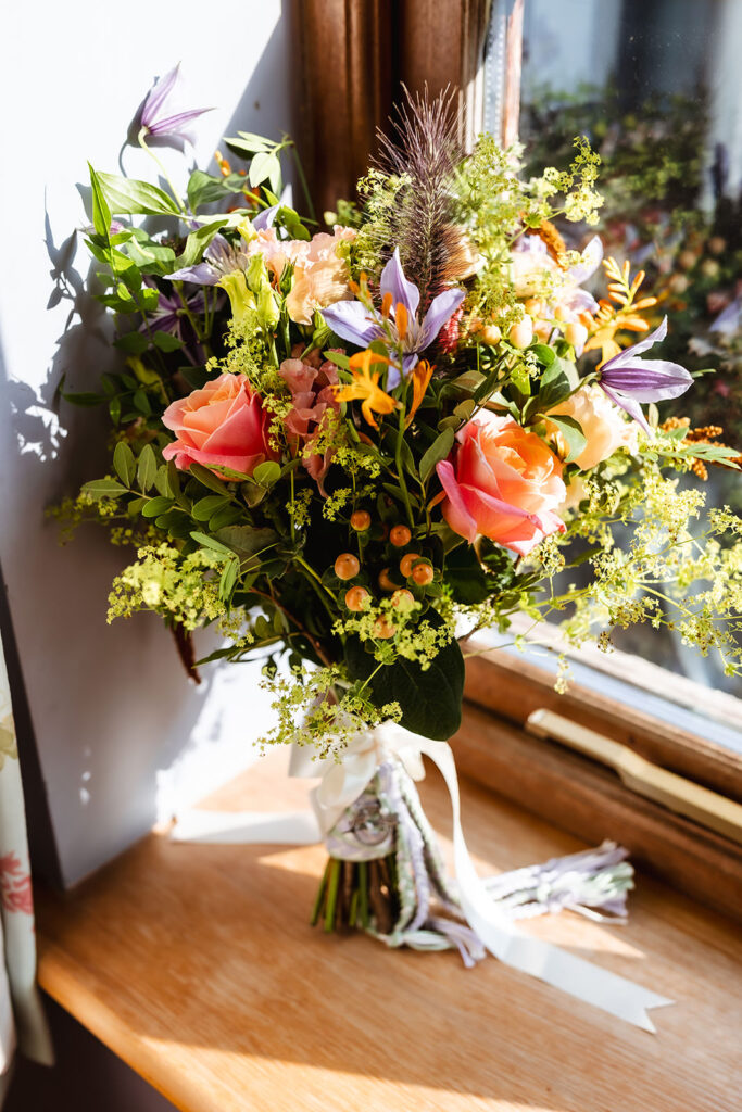 A vibrant bouquet of pink roses, purple lilies, yellow flowers, and green foliage sits on a wooden windowsill, sunlight streaming through the window and illuminating the colorful arrangement.
