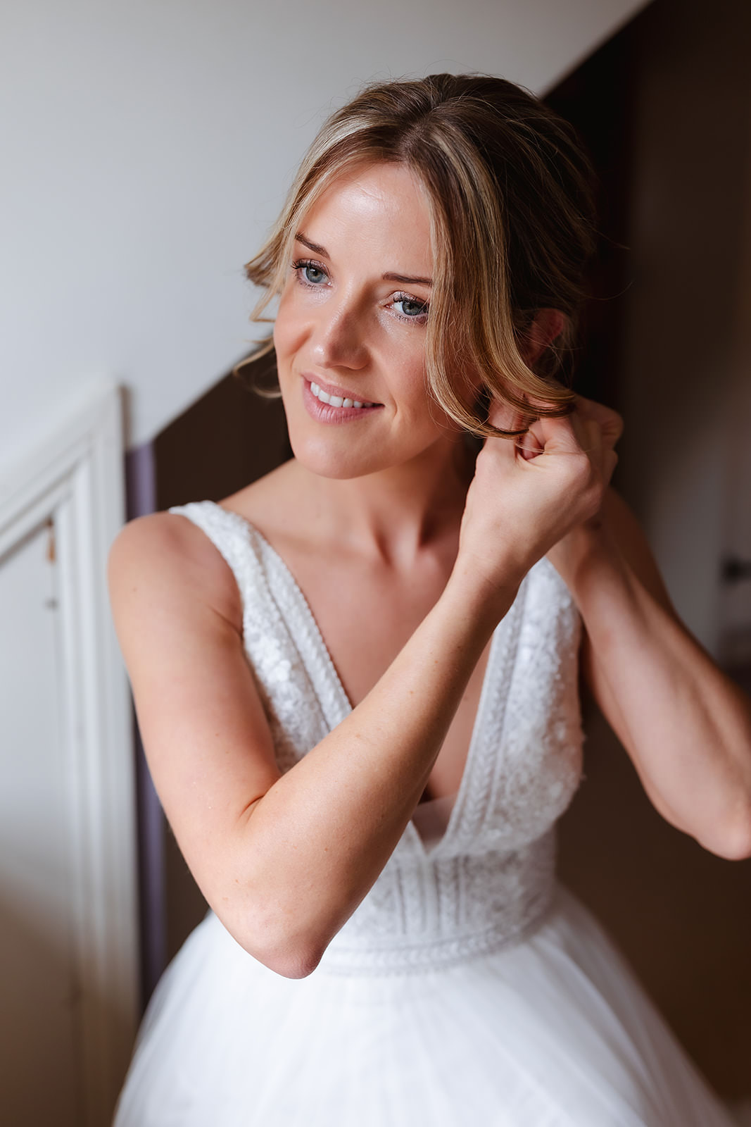 A smiling bride in a white wedding dress adjusts an earring, standing indoors near a white wall with soft natural lighting.