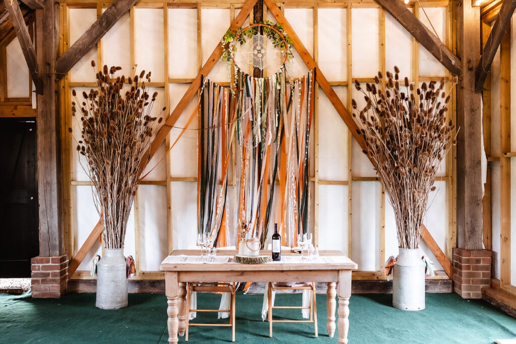A rustic wooden table with two chairs is set with glassware and a wine bottle, framed by tall vases of dried foliage and colorful ribbon streamers hanging on a barn wall.