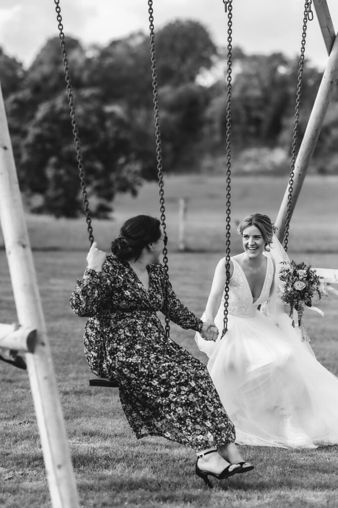 Two women are sitting on swings in a grassy outdoor setting. One wears a floral dress, and the other, a bride in a white gown holding a bouquet, smiles and looks at her companion. The scene is joyful and candid.