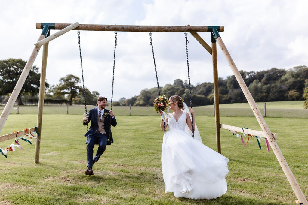 A bride in a white dress stands next to a groom on a swing in a grassy field, both smiling and holding hands. Colorful ribbons decorate the swing set, with trees and a fence in the background.