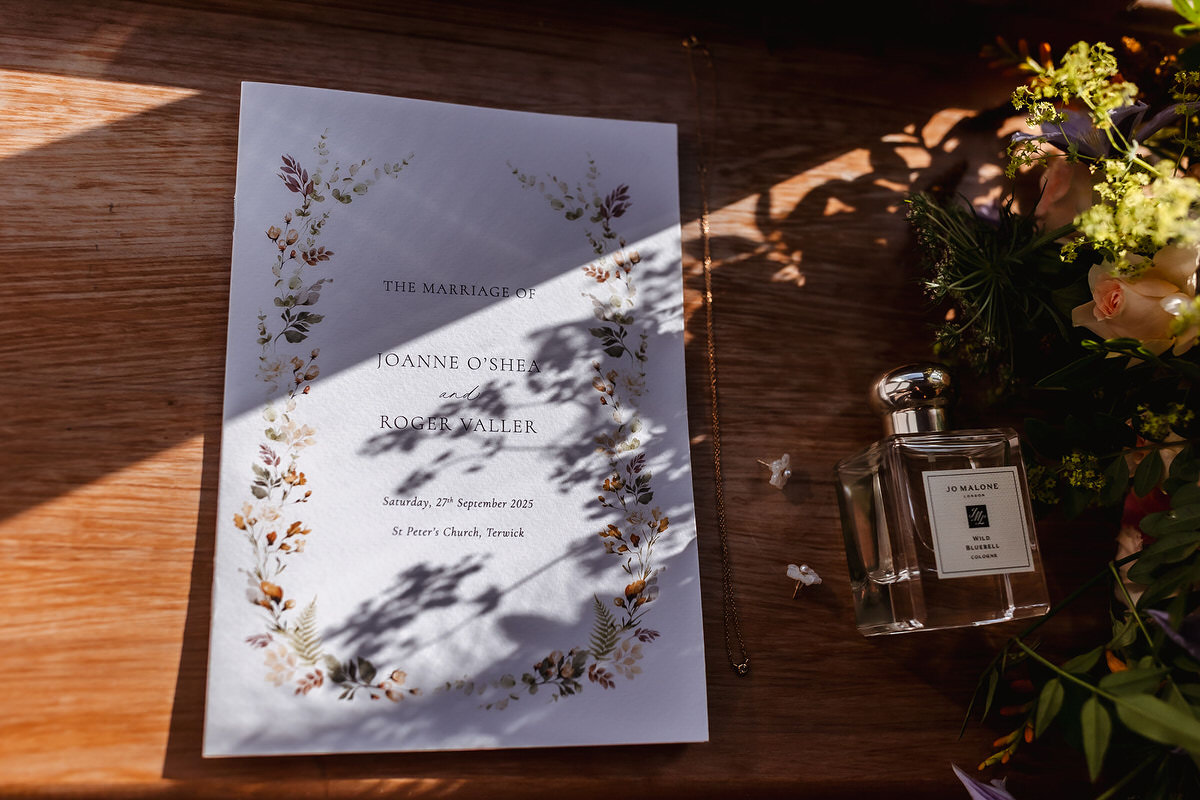 A wedding invitation for Joanne O’Shea and Roger Waller lies on a wooden surface, decorated with floral borders. Next to it is a Jo Malone perfume bottle and some flowers, with shadows falling across the scene.