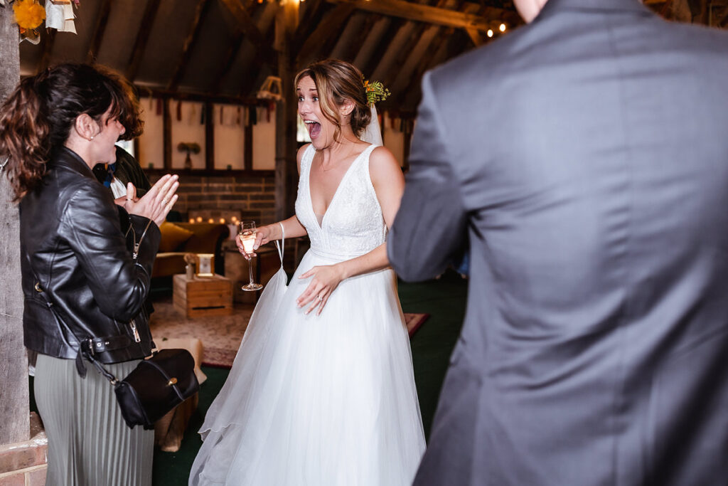 A bride in a white dress stands indoors, looking surprised and delighted, holding a glass of champagne. A woman in a leather jacket claps excitedly nearby, while a man in a suit stands in the foreground.