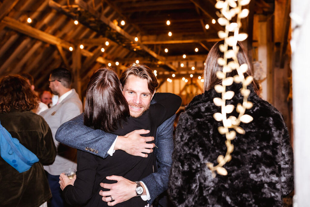 A man in a suit smiles while hugging a woman at a warmly lit indoor gathering with string lights and wooden beams visible. Other people are mingling in the background.